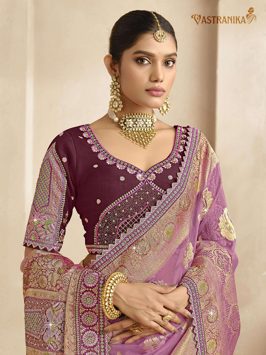 Woman wearing a traditional purple saree with intricate patterns and gold jewelry against a beige background.