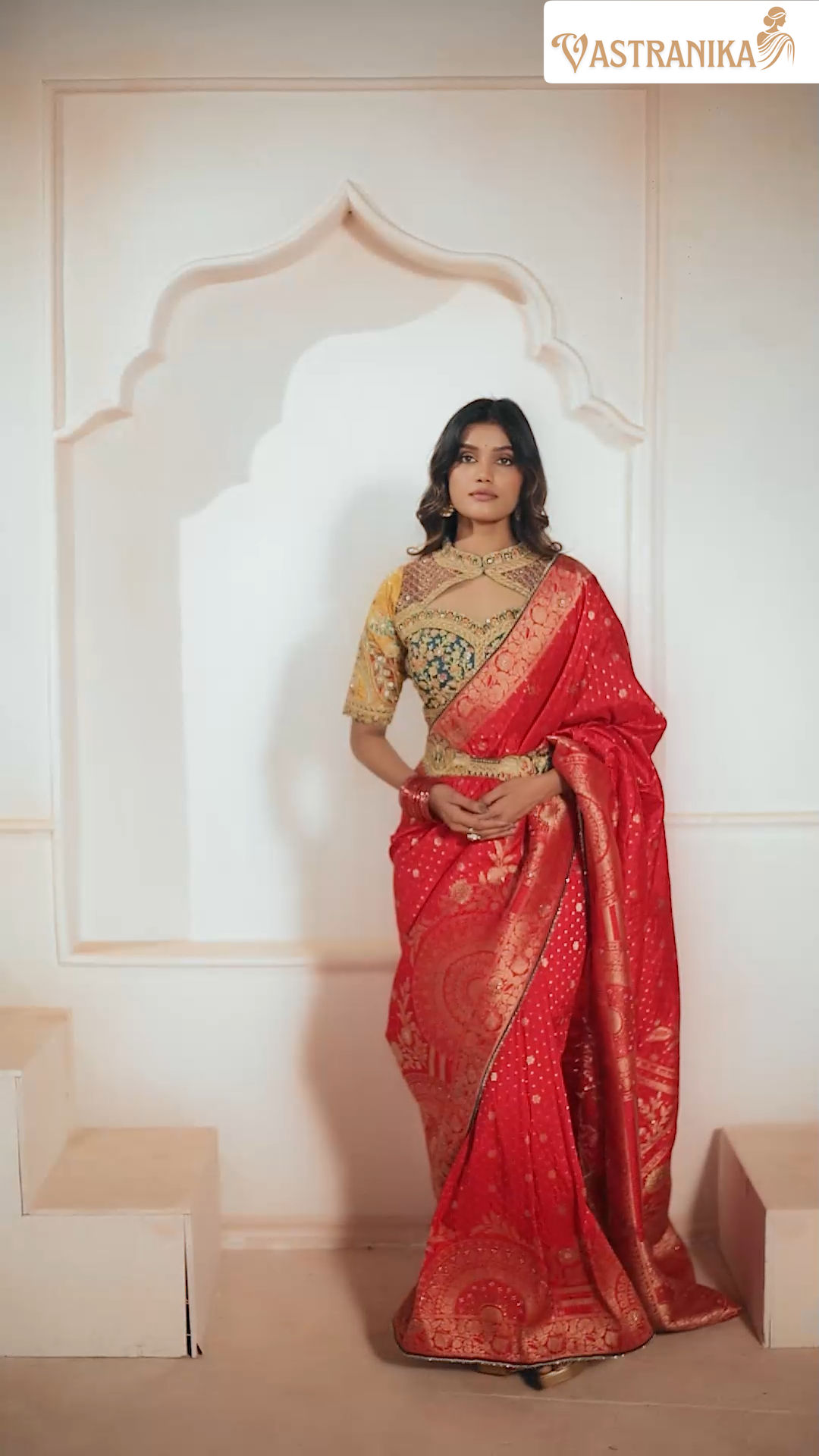 Woman in a red saree with gold patterns standing against a white wall.